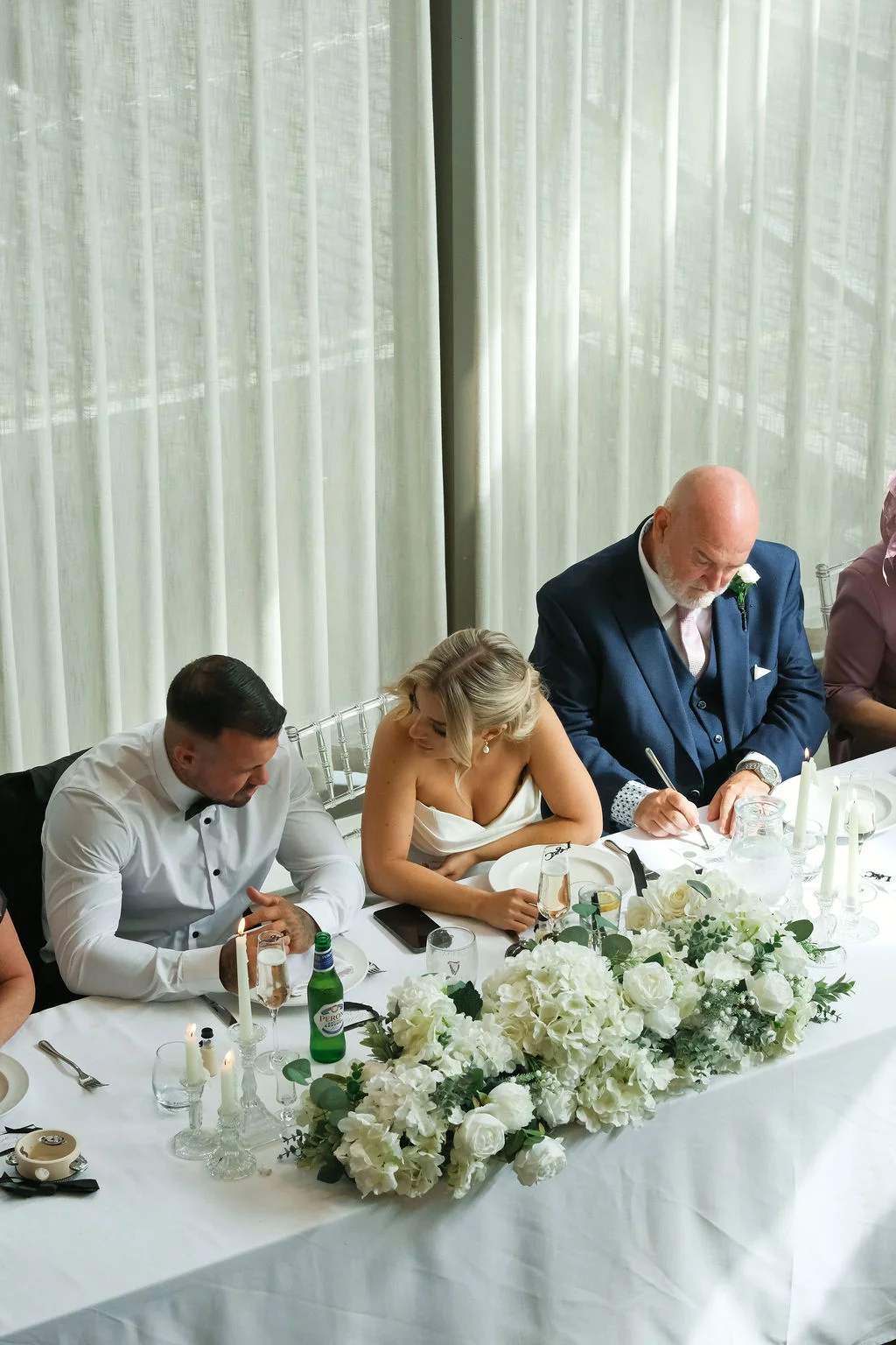 The bride, groom, and father of the bride sit together at a beautifully decorated top table adorned with white hydrangeas, roses, and glowing candles while someone signs a document.