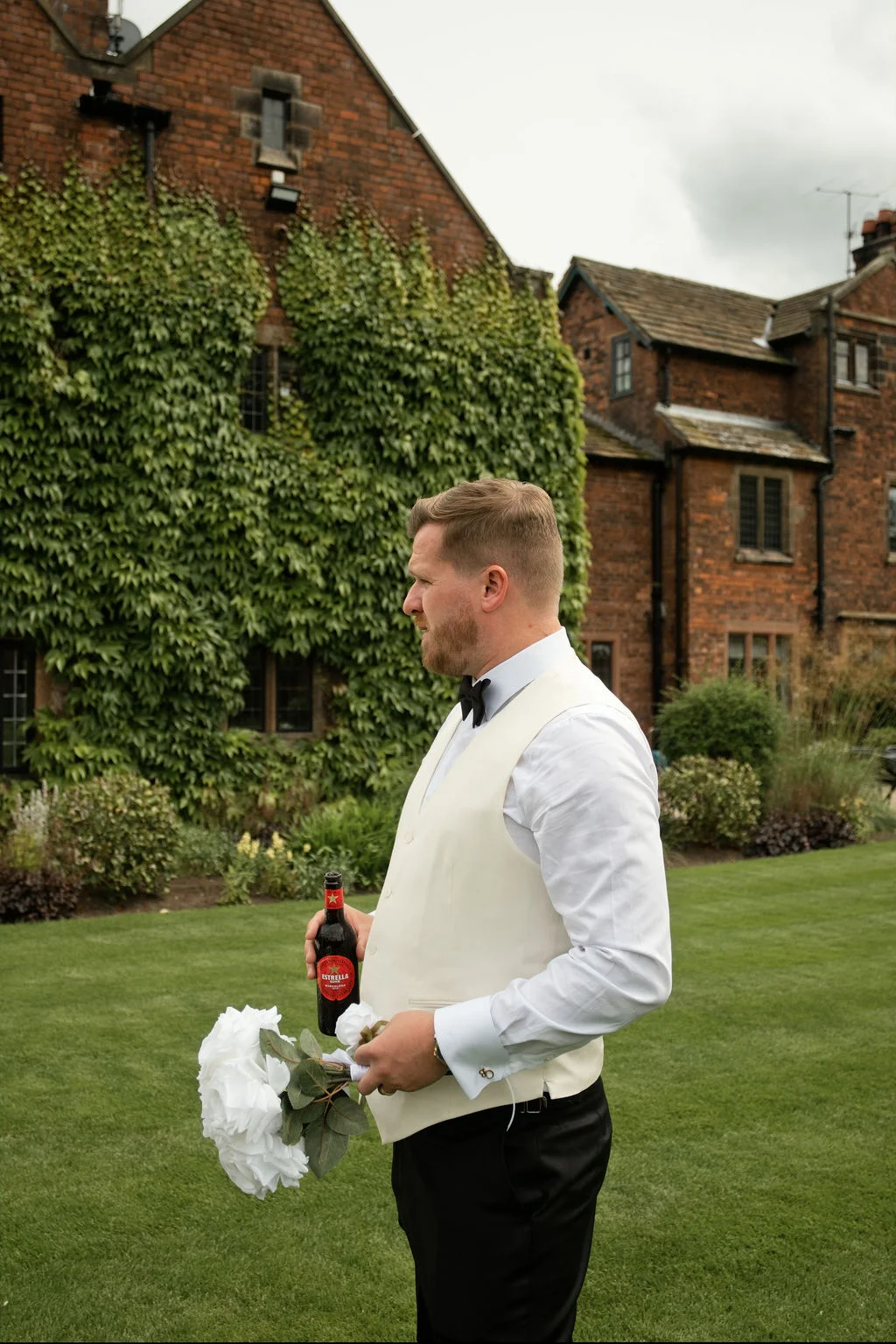 A groom stands outdoors on a lawn in front of an ivy-covered brick building, holding a bottle of beer and a white floral bouquet.