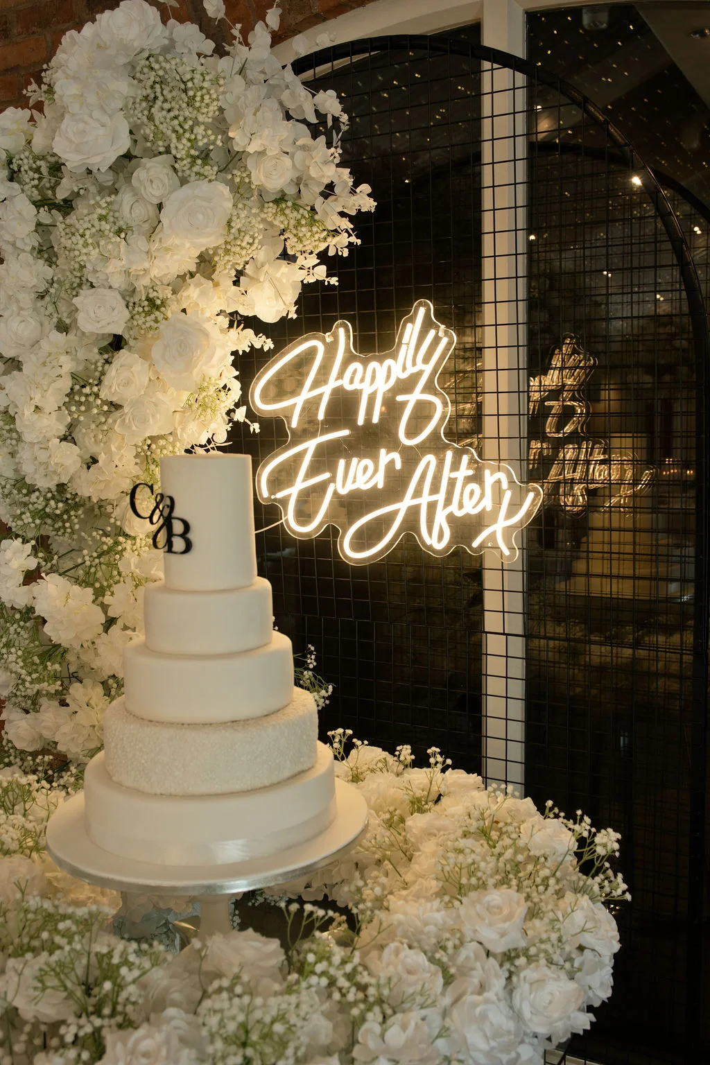 A magnificent tall white tiered wedding cake sits surrounded by lush white roses and baby's breath, positioned in front of a modern dark grid backdrop featuring an illuminated neon sign.