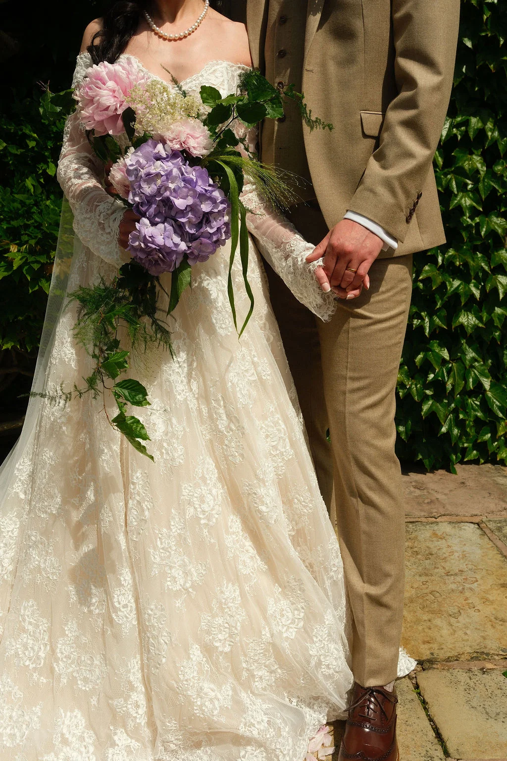 A newlywed couple holds hands outdoors against an ivy-covered wall, with the bride wearing an off-the-shoulder lace gown and holding a cascading bouquet of pink peonies and purple hydrangeas, while the groom wears a tan tweed suit.