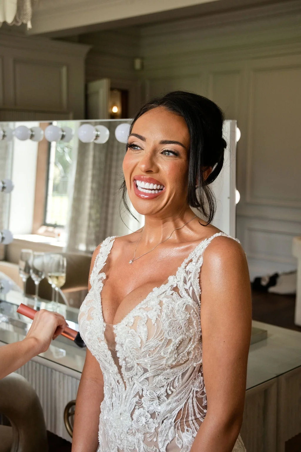 A glowing bride smiles broadly as she finishes getting ready in a bright room, wearing a lace wedding dress with a plunging neckline and her dark hair styled in an elegant updo.