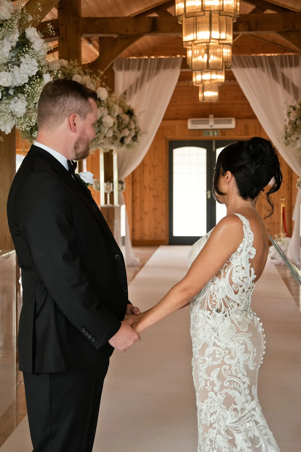 A newlywed couple holds hands at the altar inside a rustic oak-framed barn featuring a white aisle runner and elegant chandeliers.