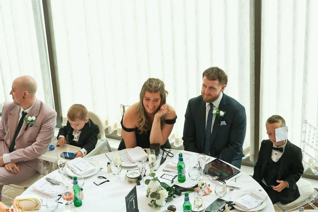 Guests and family members sit at a brightly lit round wedding breakfast table, smiling and listening during the speeches.