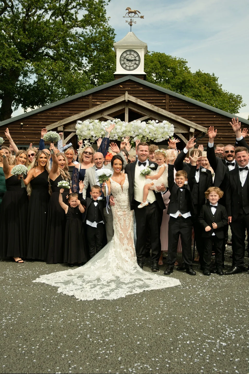 A large wedding party and guests celebrate together outside a rustic wooden barn with a clock tower, cheering with their arms raised as confetti scatters on the ground.