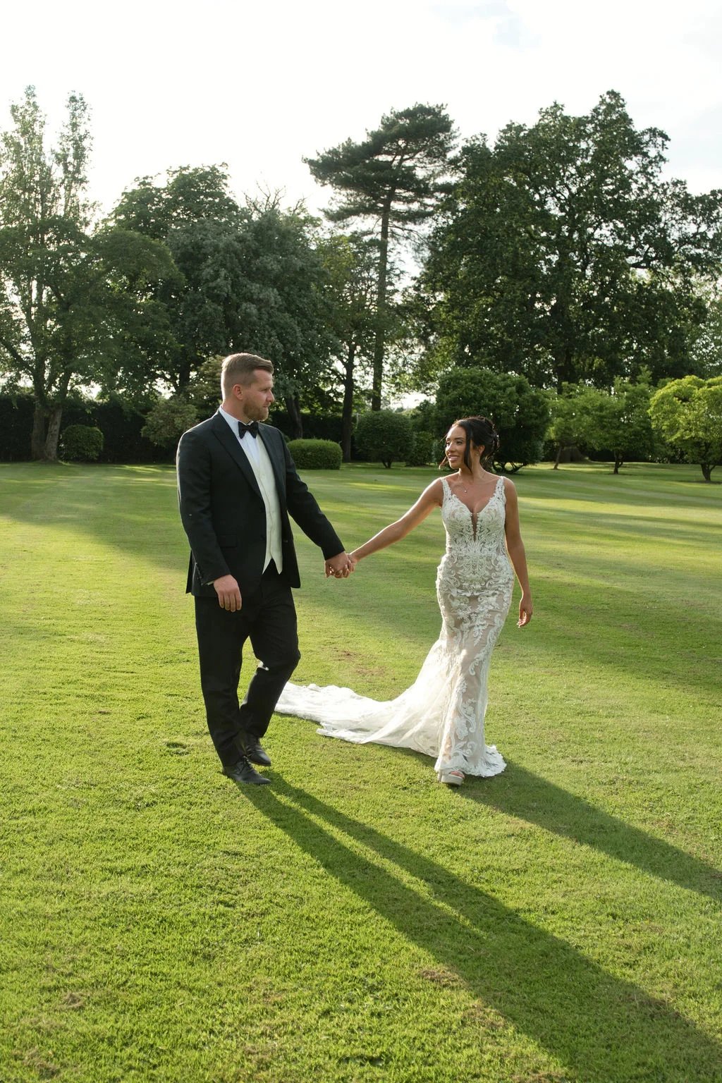 A bride and groom walk hand in hand across a sunlit green lawn, sharing a loving glance during their outdoor couples portrait session.