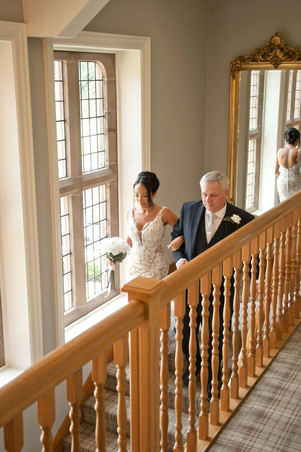 A bride holding a white rose bouquet walks down a wooden staircase alongside her father inside a manor house, captured in soft natural light.