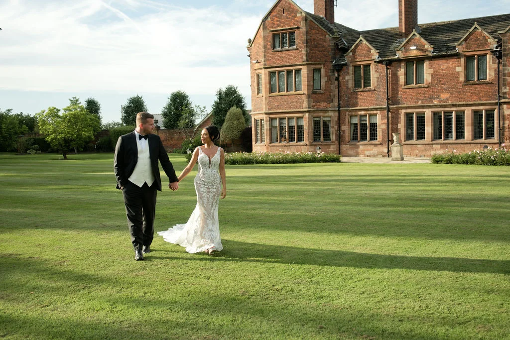 A bride in a lace mermaid gown and a groom in a black tuxedo hold hands while walking across the manicured lawn of a historic country house.
