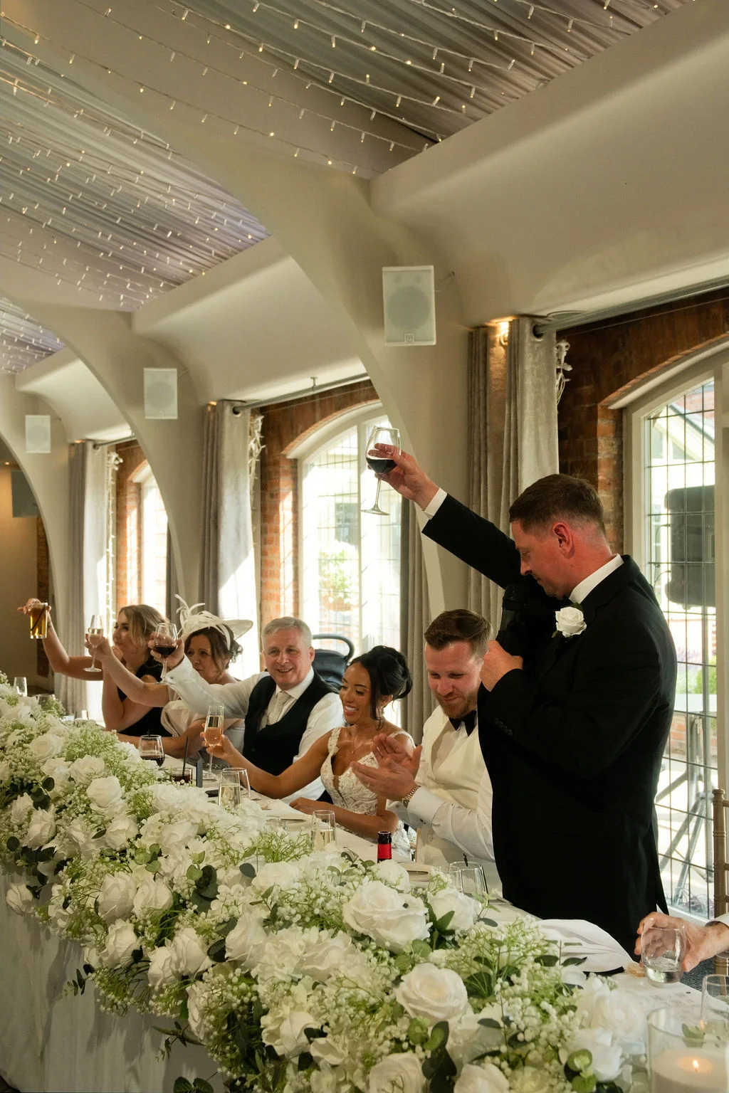 The groom raises a glass to toast the guests during the wedding speeches at a long white-floral decorated top table.
