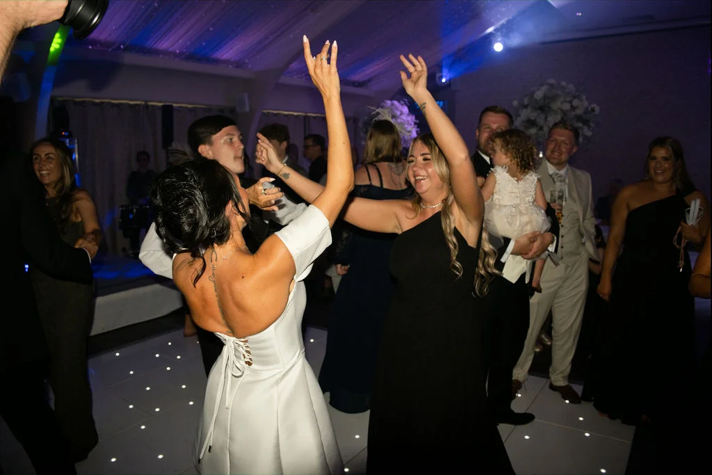A bride in a white reception dress and guests with their hands raised dance joyfully on an illuminated dance floor during a lively wedding evening party.