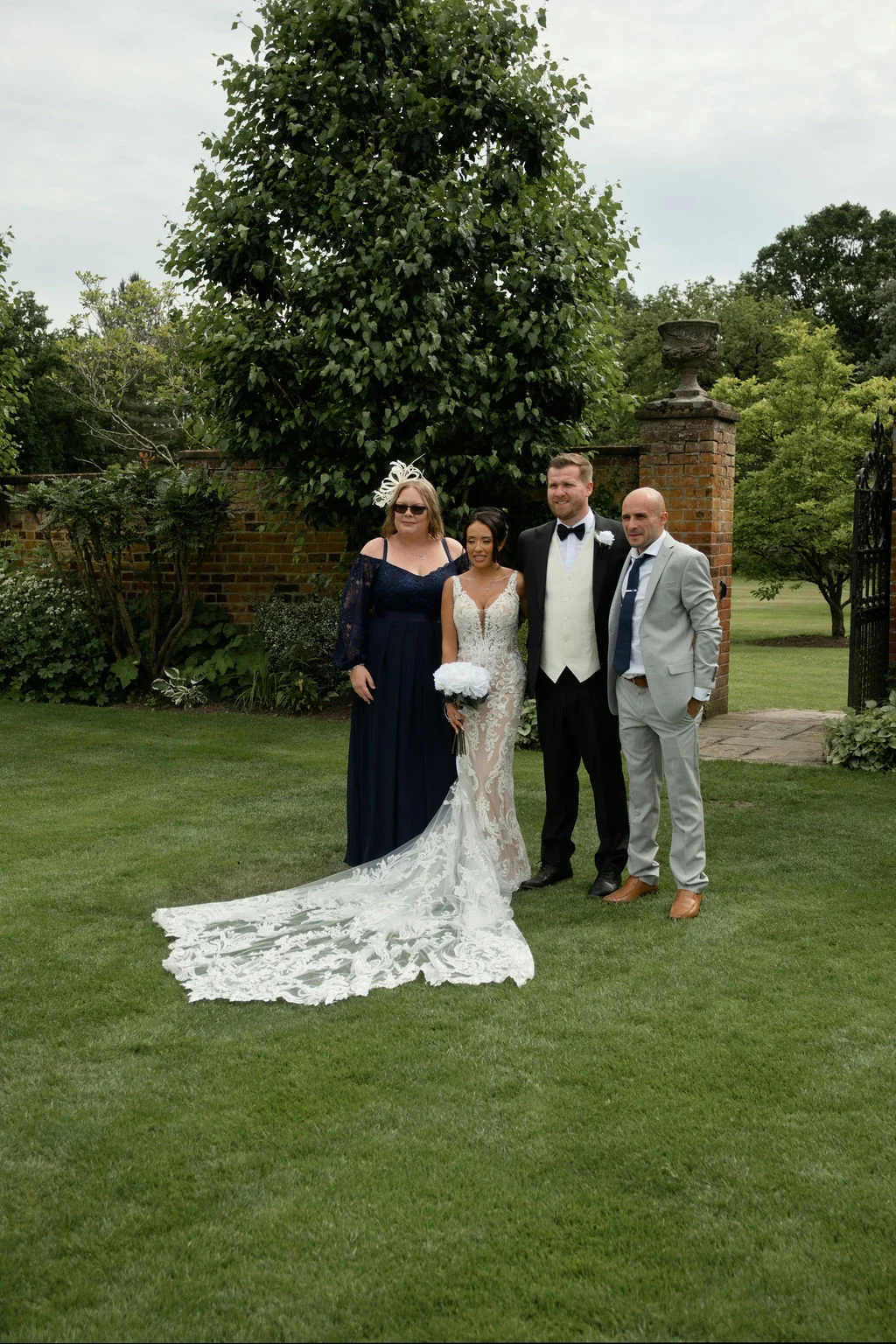 A bridal party posed outdoors on a manicured lawn next to a brick wall and stone pillar, featuring a bride in a lace gown with a long train, a groom in a tuxedo, and family members.