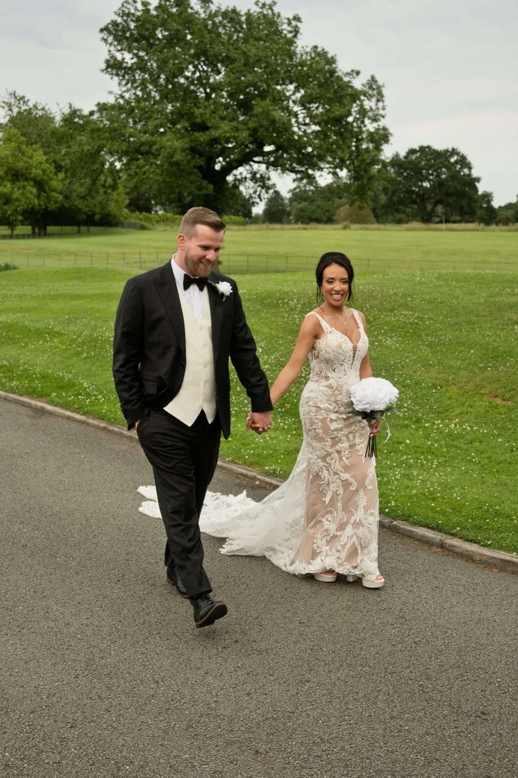 A newlywed couple happily walks hand in hand along a paved path bordered by green lawns and rolling countryside.