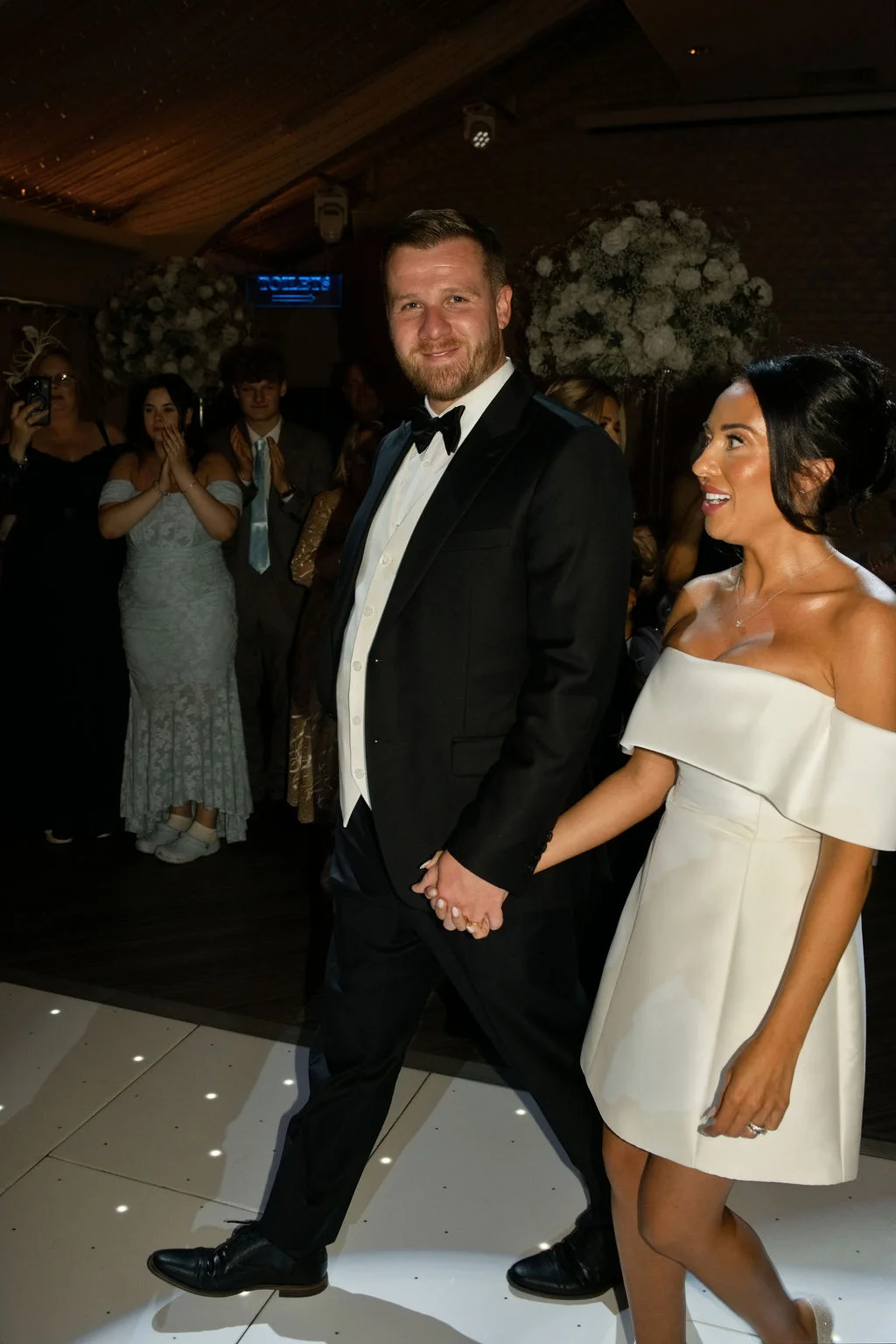Newlyweds make their entrance into the evening reception hand in hand, with the bride wearing a stylish short off-the-shoulder dress and the groom in a classic tuxedo.
