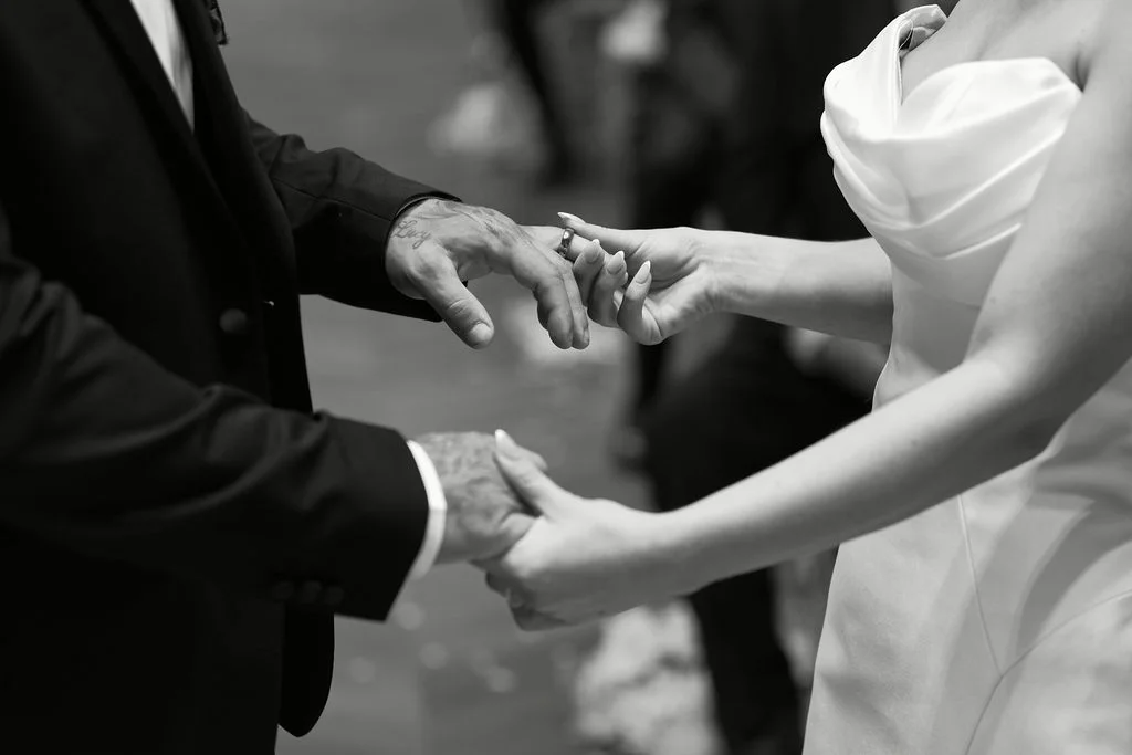 A close-up black and white shot captures the bride placing a wedding ring onto the groom's hand during the ceremony.