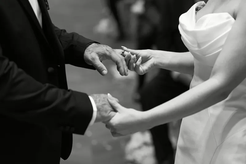 A close-up black and white shot captures the bride placing a wedding ring onto the groom's hand during the ceremony.