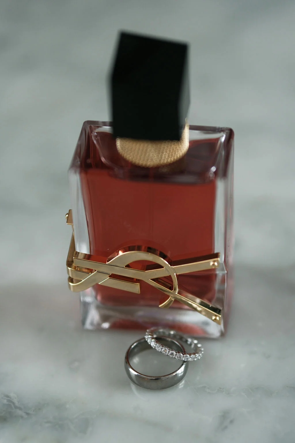 A close-up bridal detail shot features a luxury perfume bottle resting on a marble surface alongside a diamond eternity band and a plain metal wedding band.
