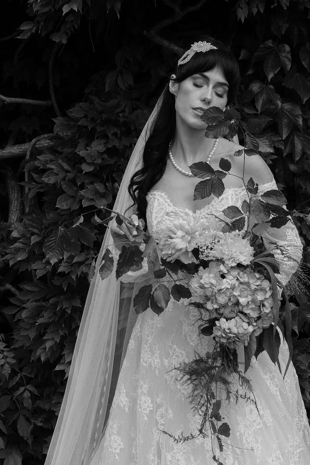 A stunning black and white portrait of a bride posing gracefully against a lush ivy-covered wall, holding a textured floral bouquet and wearing an off-the-shoulder lace gown.