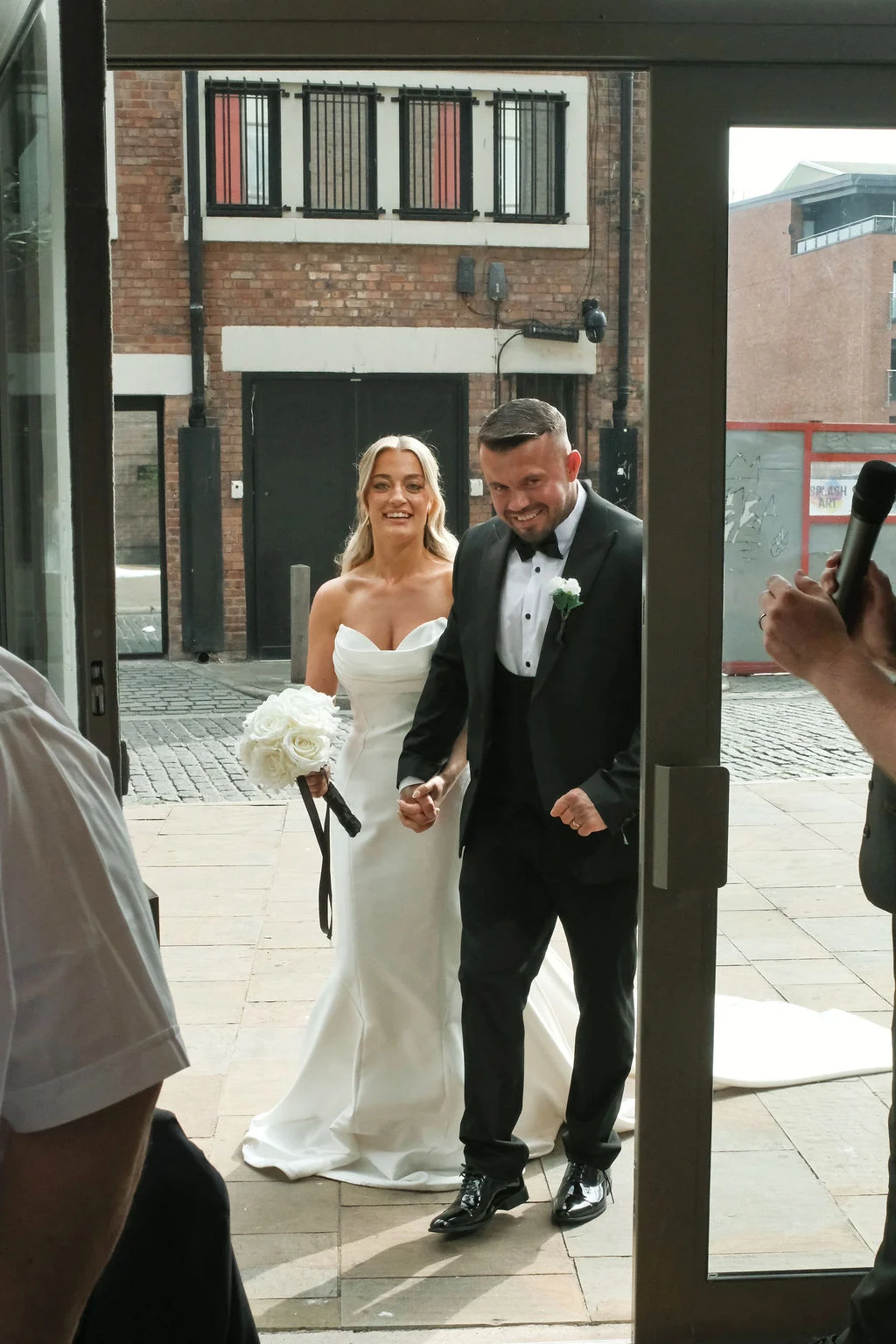 A newlywed couple walks hand in hand through glass doors onto an urban stone courtyard, with the smiling bride holding a bouquet of white roses.