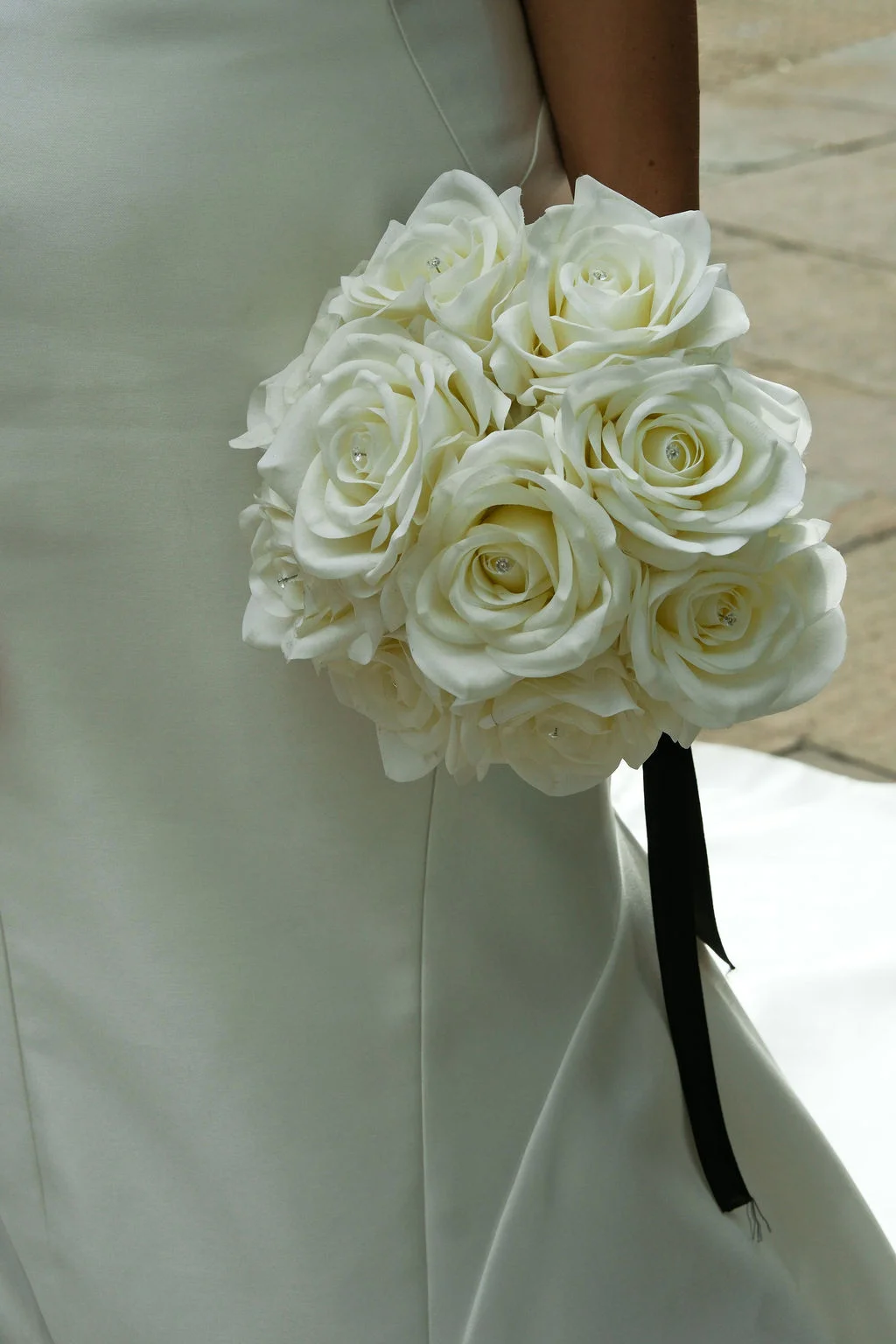 A bride holding a compact round bouquet of pristine white roses tied with a dark ribbon against a simple satin wedding dress.