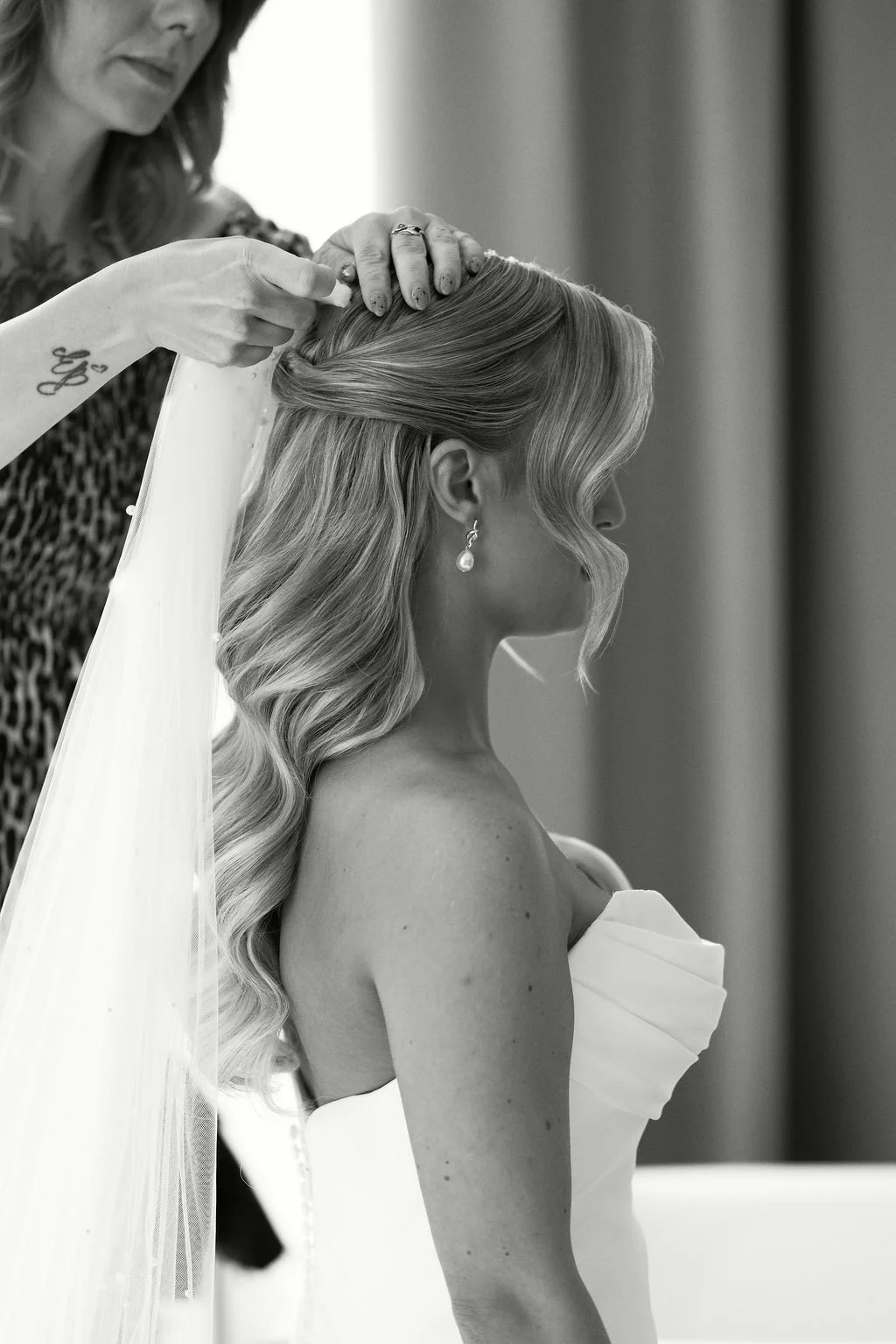 A bridal stylist delicately fastens a veil into the blonde hair of a bride wearing a strapless gown during morning preparations.