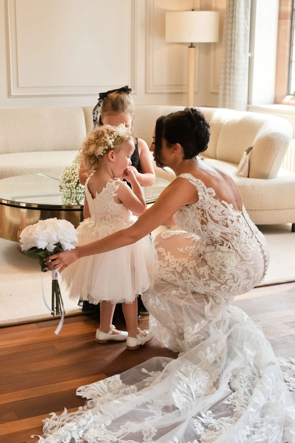 A bride in a fitted lace wedding dress with a dramatic train kneels on a wooden floor to show her bouquet to a young flower girl wearing a white tulle dress.