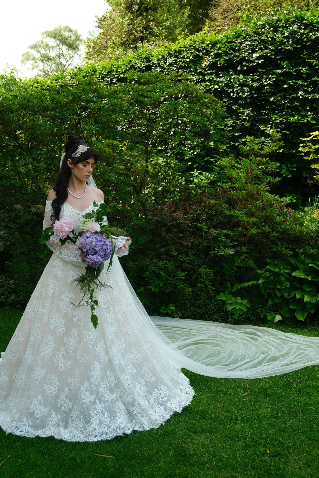 A stunning bride in an off-the-shoulder lace a-line gown holds a cascading bouquet of peonies and hydrangeas in a lush garden setting with her long veil trailing on the grass.