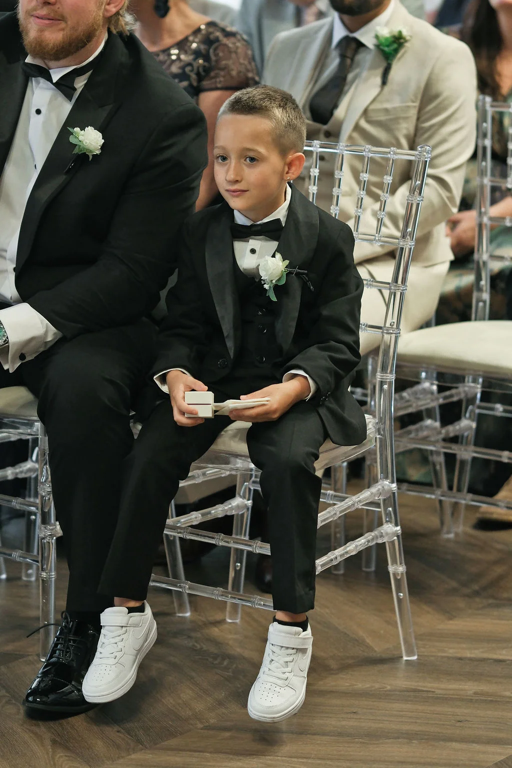 A young page boy sits in a clear ghost chair during a wedding ceremony, wearing a smart black tuxedo and white trainers while holding a ring box.