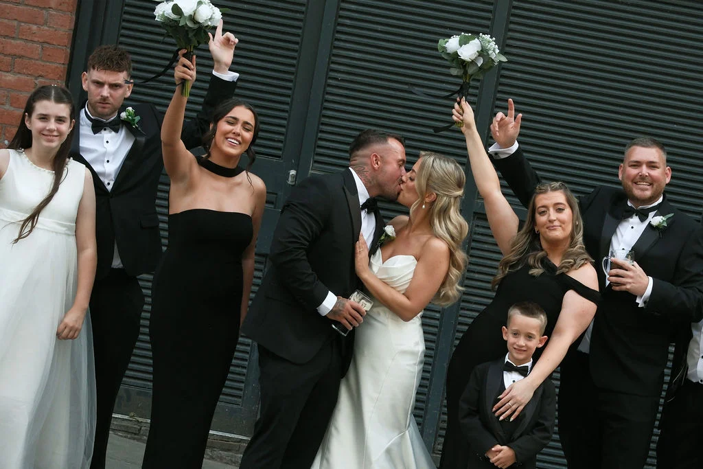 A newlywed couple share a kiss in the centre of their wedding party, with bridesmaids in black dresses and groomsmen in black tuxedos outside an industrial building.
