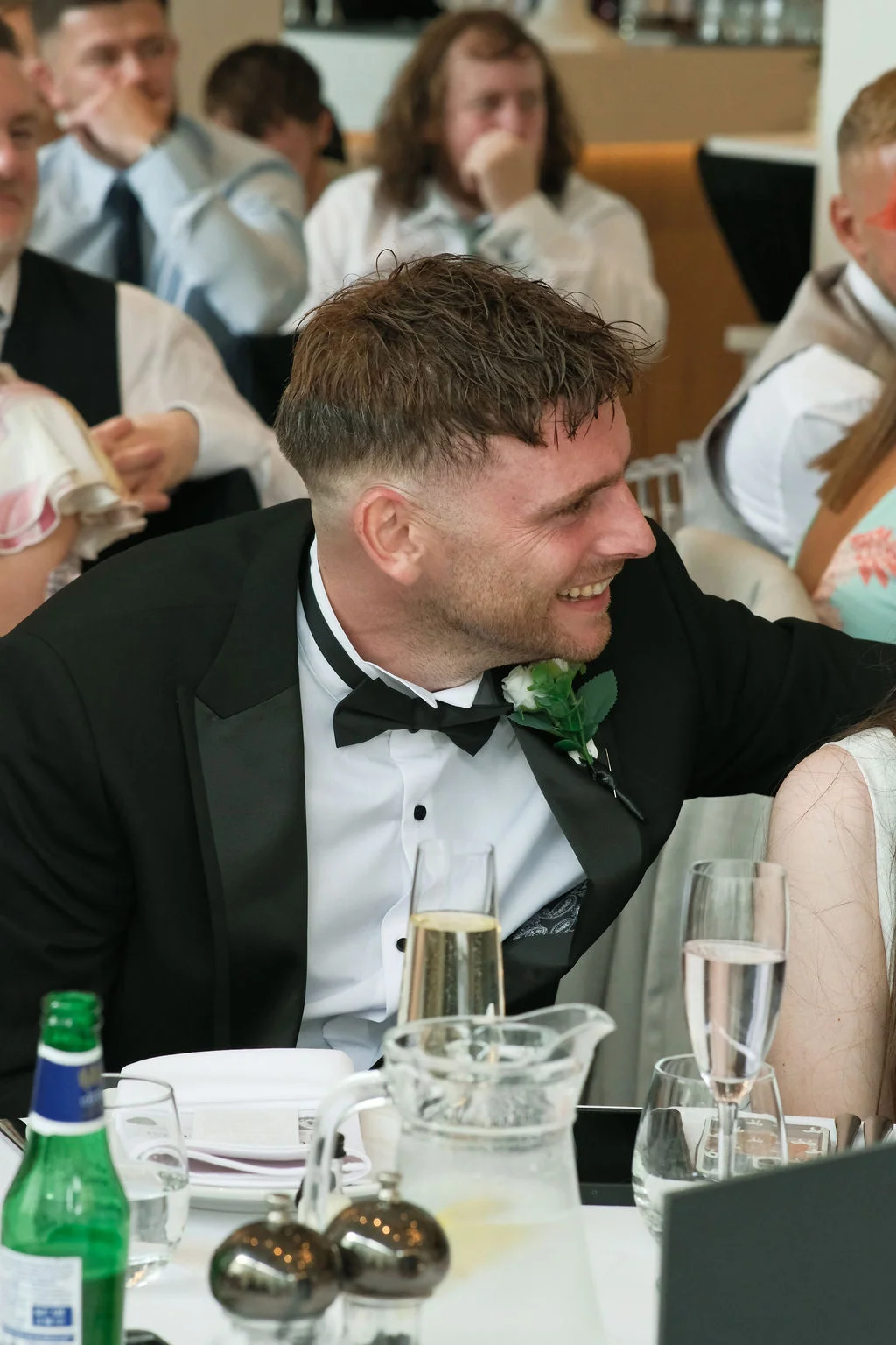 A smiling groom wearing a black tuxedo and bow tie laughs during the wedding reception speeches.