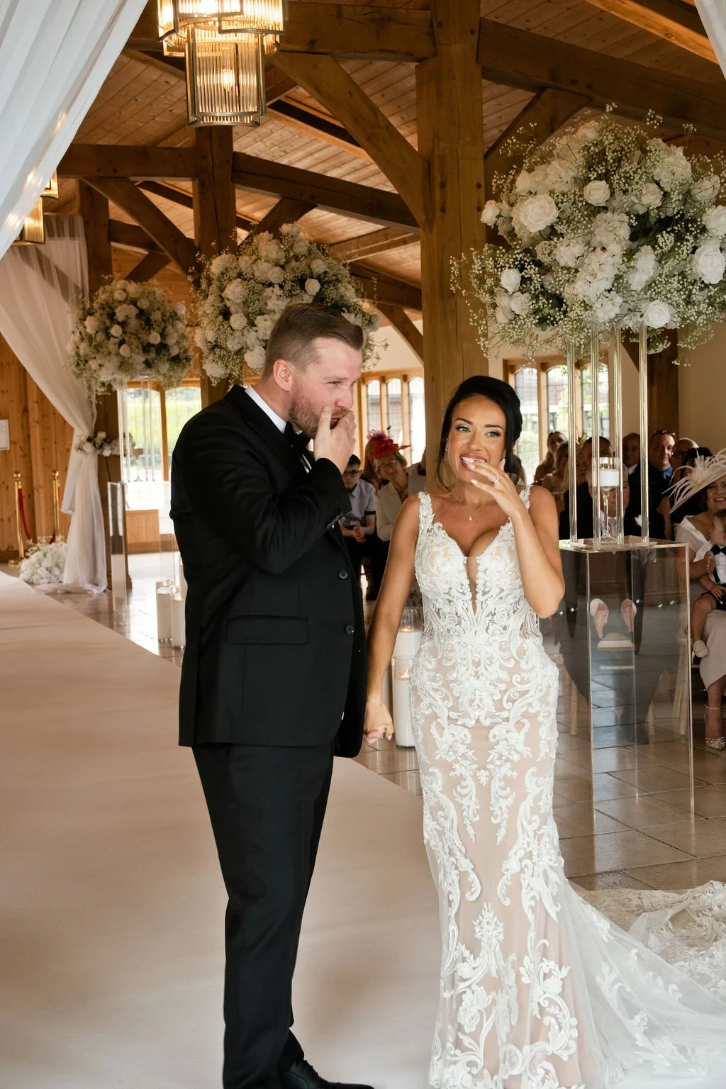A newlywed bride and groom laugh together hand in hand at the top of the aisle inside an oak framed barn venue decorated with white floral pedestals.