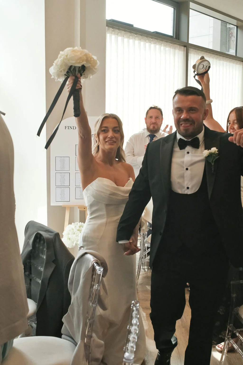 A newlywed couple excitedly enters their wedding reception, holding hands as the bride raises her bouquet in the air and guests cheer in the background.