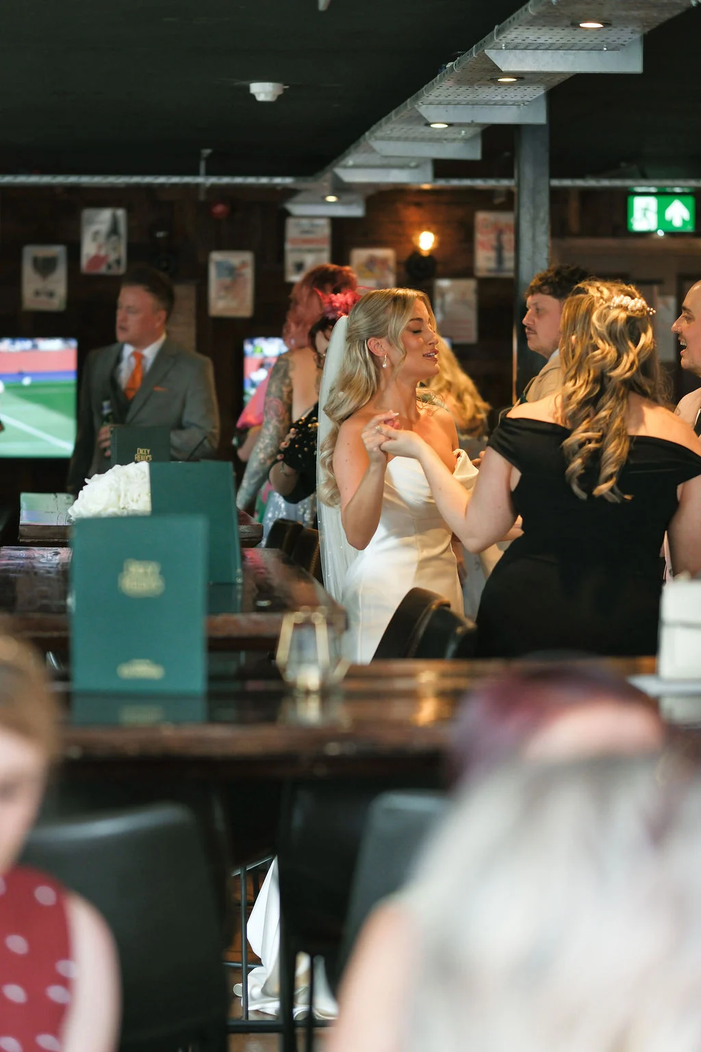 A smiling bride in a strapless satin gown dances and holds hands with a guest inside a dimly lit pub venue.