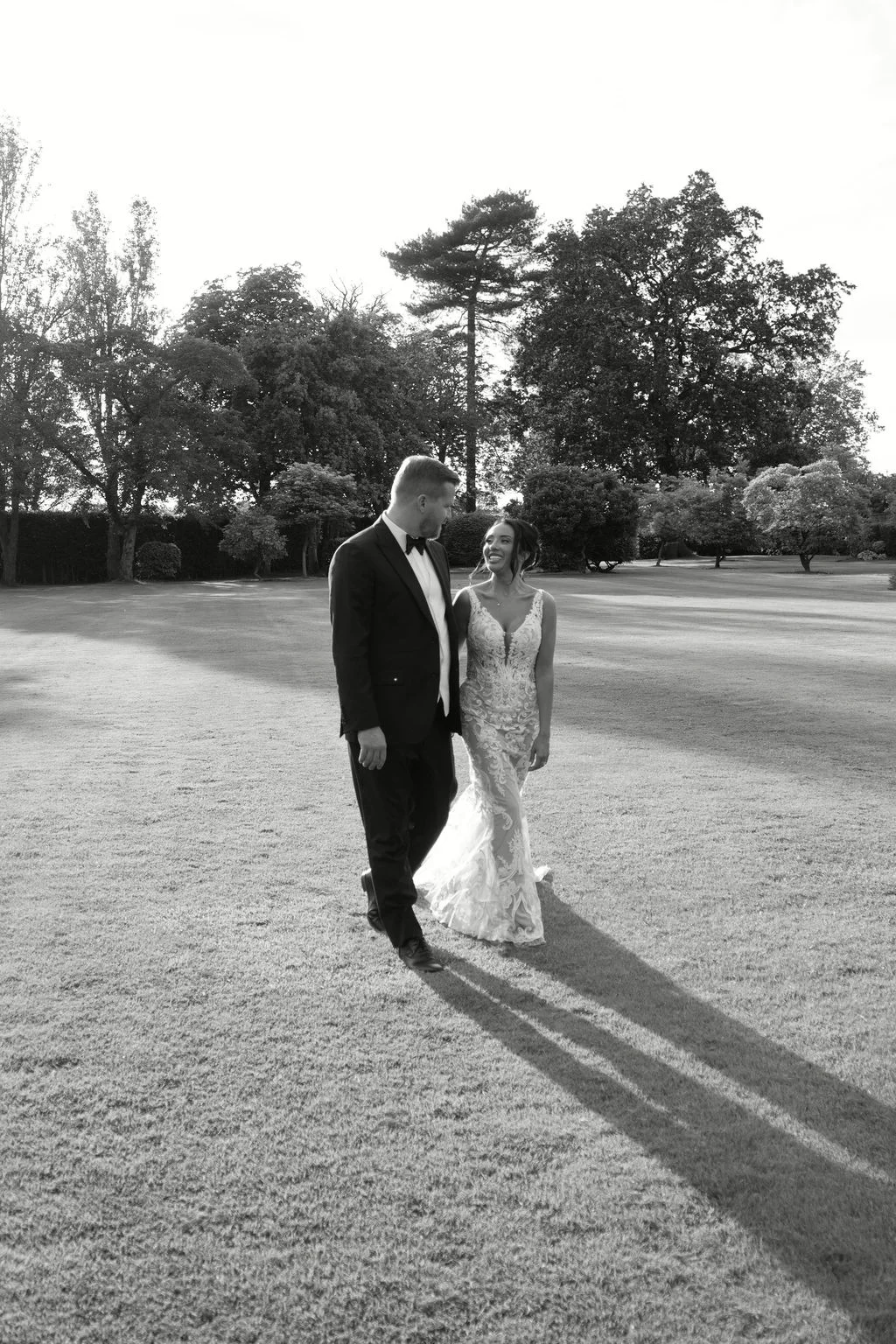 A newlywed couple strolls across a sunlit manicured lawn in a timeless black and white photograph, casting long shadows behind them.