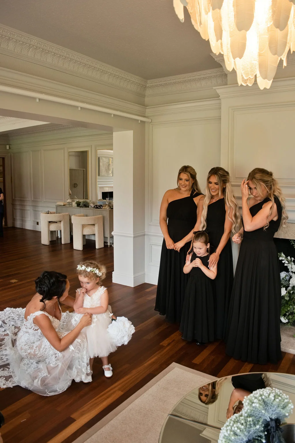 A bride in a lace gown kneels to tend to a little flower girl in a white dress, while three smiling bridesmaids in black dresses look on inside an elegant room.