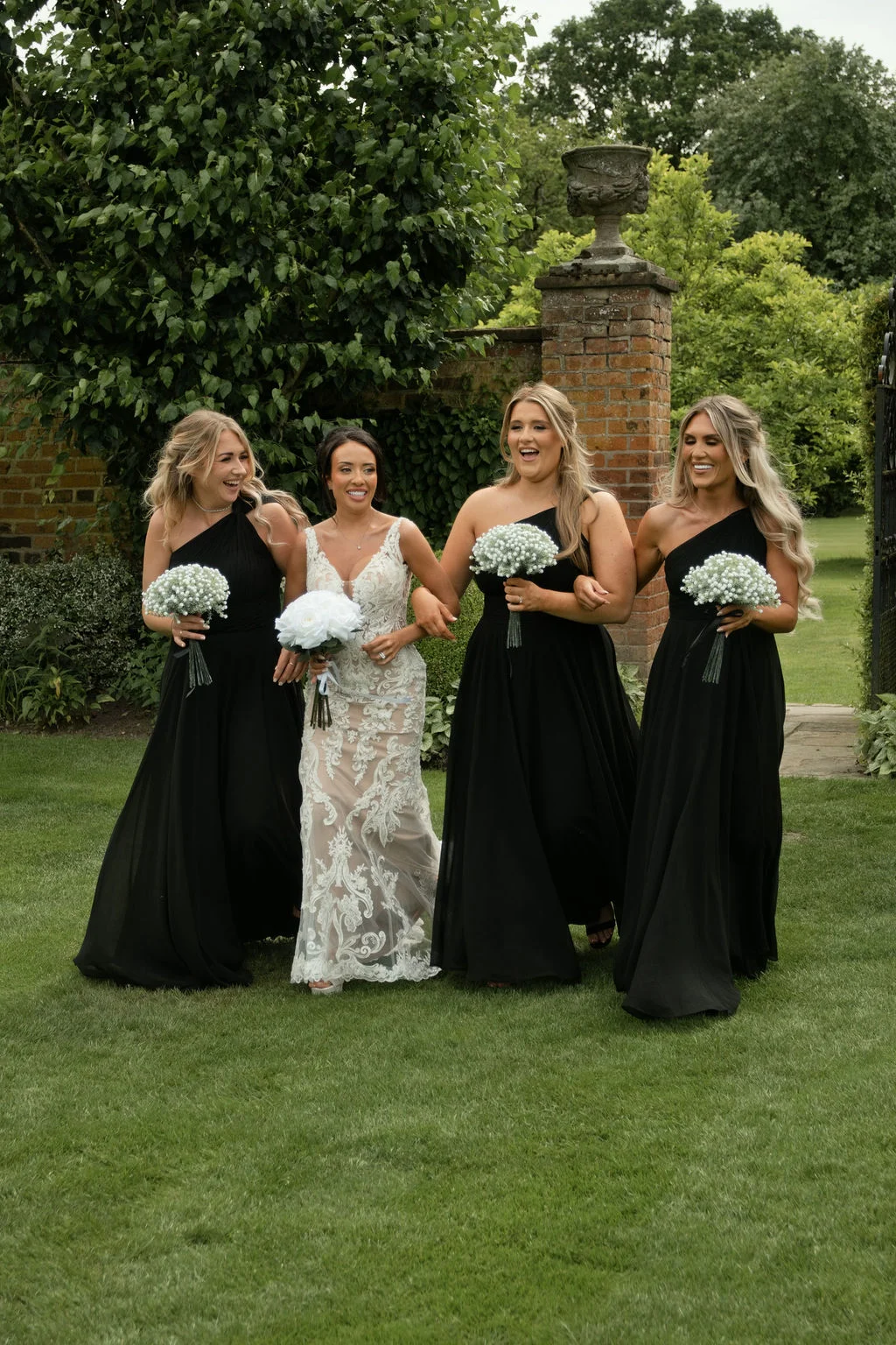 A joyful bride in an intricate lace gown walks and laughs through a manicured garden accompanied by her bridesmaids wearing matching black one-shoulder dresses holding gypsophila bouquets.