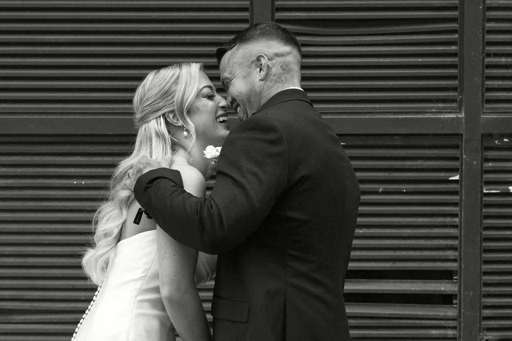 A black and white photograph of a bride and groom standing nose-to-nose against a slatted background, laughing joyfully together.