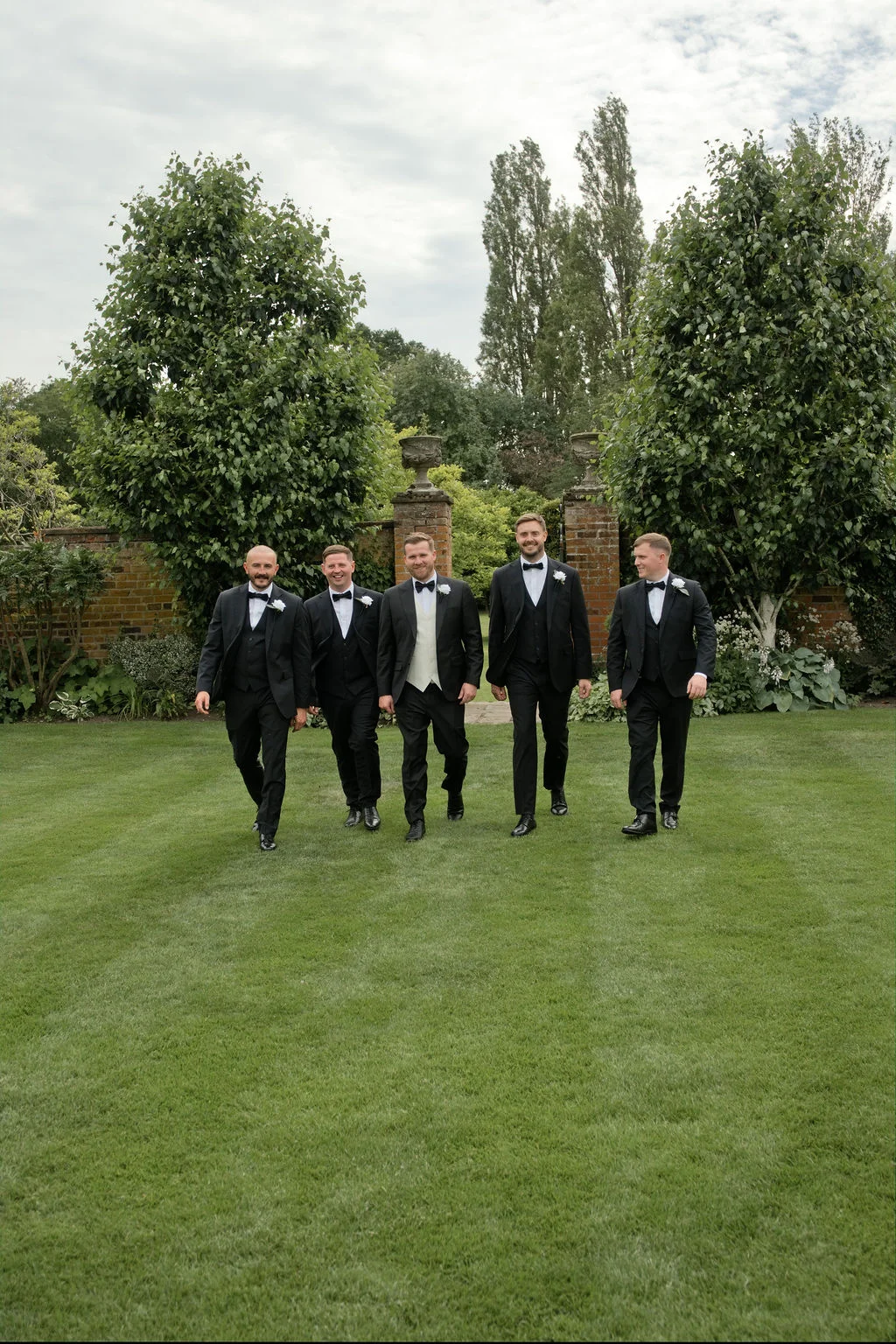 Five groomsmen in matching black tuxedos and bow ties walk together across a manicured lawn in front of an English country garden wall.