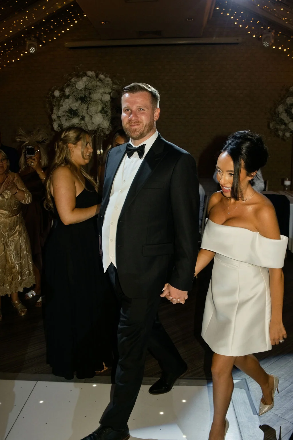 A smiling newlywed couple walks hand-in-hand across an illuminated dance floor, with the groom in a classic black tuxedo and the bride wearing a stylish short off-the-shoulder white dress.