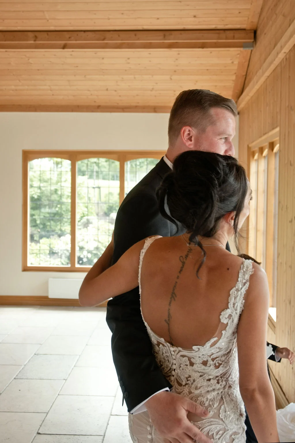 A bride in a lace dress with a low back and the groom in a dark suit embracing inside a wooden-beamed room with large windows.