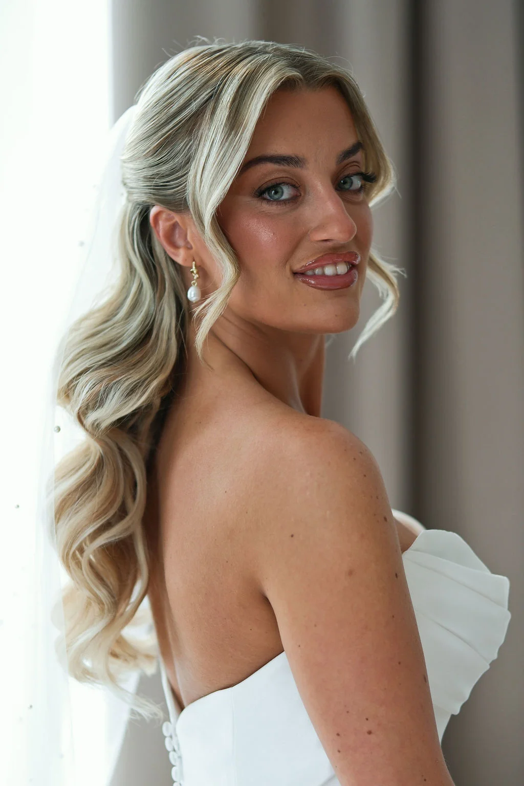 A stunning bride looking over her shoulder with a radiant smile, showcasing her half-up half-down wavy blonde hair, pearl drop earrings, and a strapless ruched wedding dress near a bright window.