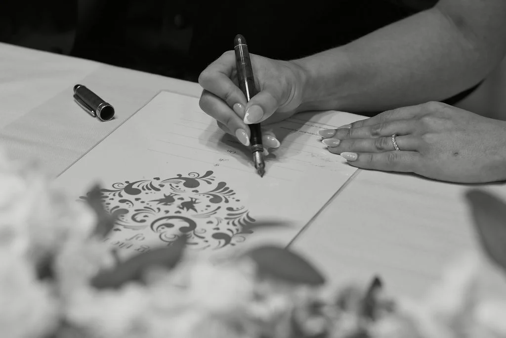 A close-up black and white photograph captures hands signing the marriage certificate during the wedding ceremony.