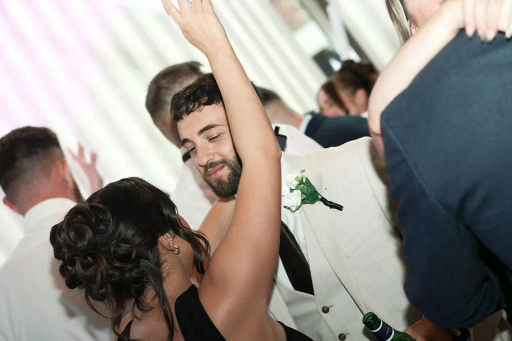 A groom in a light beige suit with a buttonhole smiles while dancing on a packed evening reception dance floor with his partner.