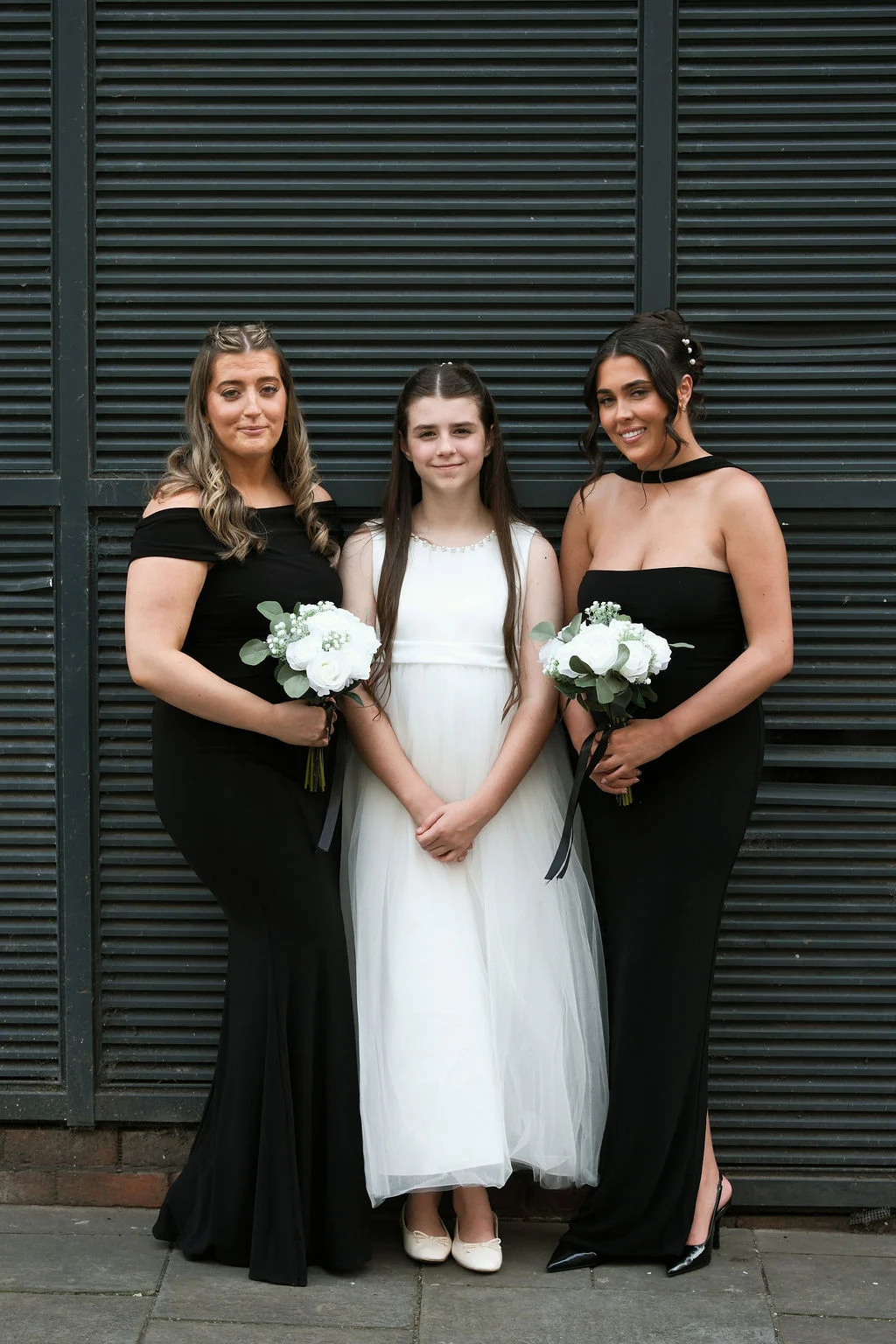 Two adult bridesmaids in elegant black evening dresses stand alongside a junior bridesmaid in a white dress, each holding bouquets of white roses against a dark slatted backdrop.