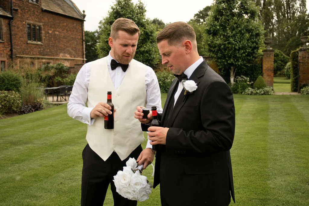Two grooms in formal wedding attire holding beer bottles and a white rose bouquet while standing on a manicured lawn outside a historic brick building.