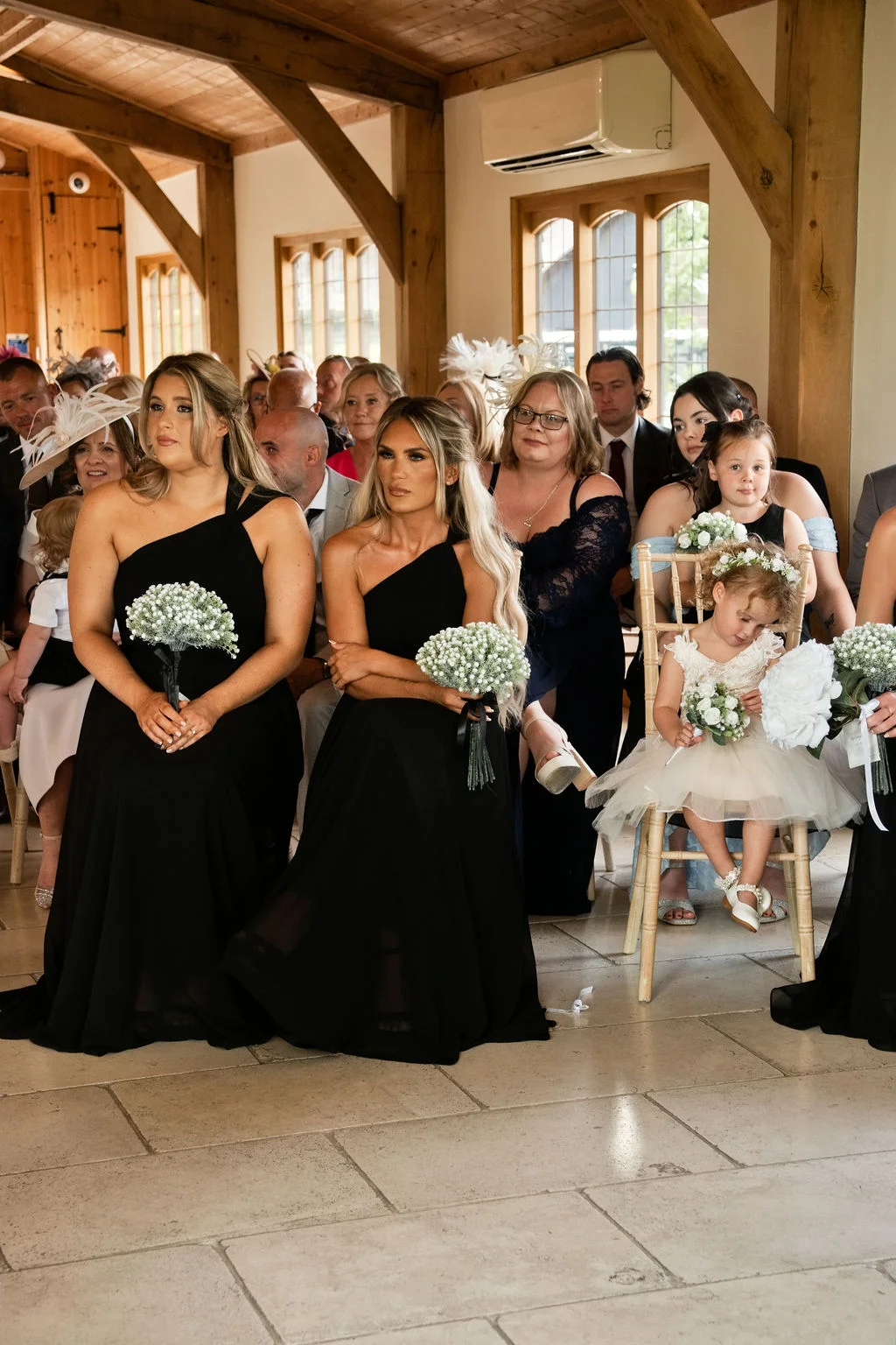 Bridesmaids wearing elegant black one-shoulder dresses and holding gypsophila bouquets are seated inside a rustic oak-framed barn during a wedding ceremony.