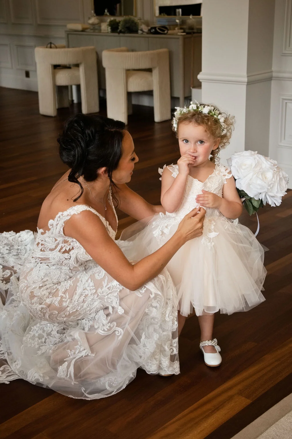 A bride in a lace gown kneels to help a flower girl in a tulle dress who holds a bouquet of white roses.