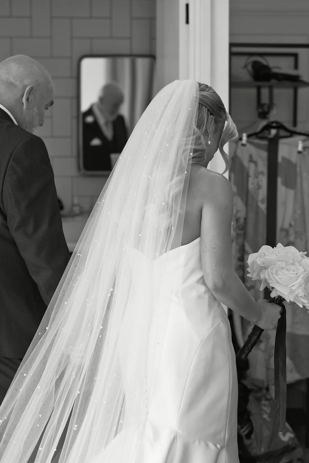 A black and white candid shot of a bride holding a rose bouquet alongside her father while wearing a satin gown and pearl-embellished veil.