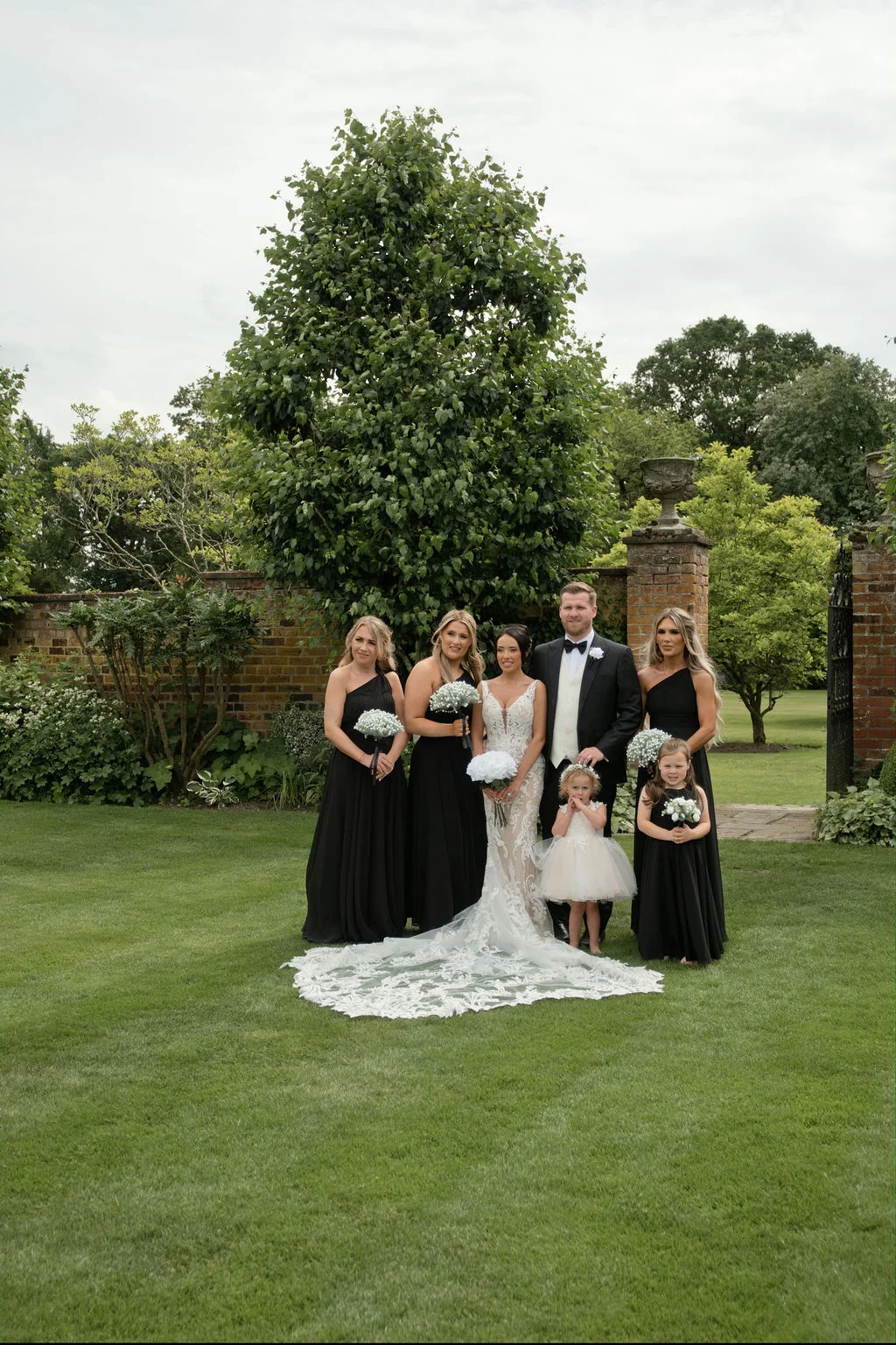 The bridal party poses on a manicured lawn in front of a walled garden, with bridesmaids in elegant black dresses flanking the bride in a lace gown with a dramatic train and the groom in a classic tuxedo.