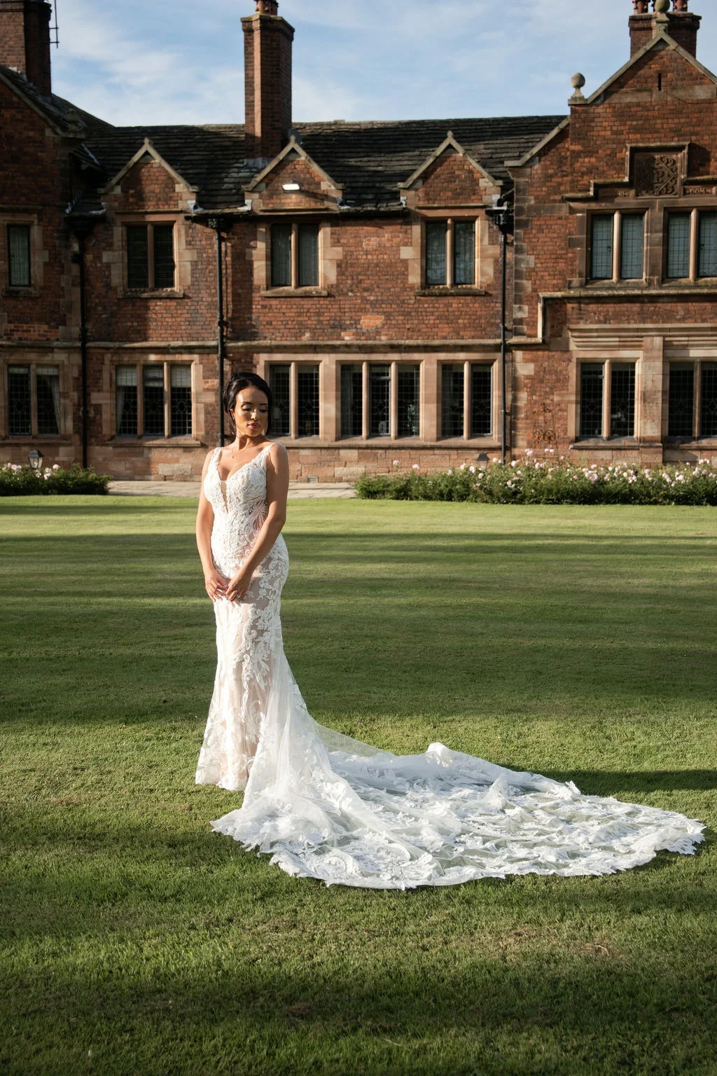 A stunning bride stands on the lawn in front of a grand red brick manor house wearing a fitted lace gown with a sweeping train.