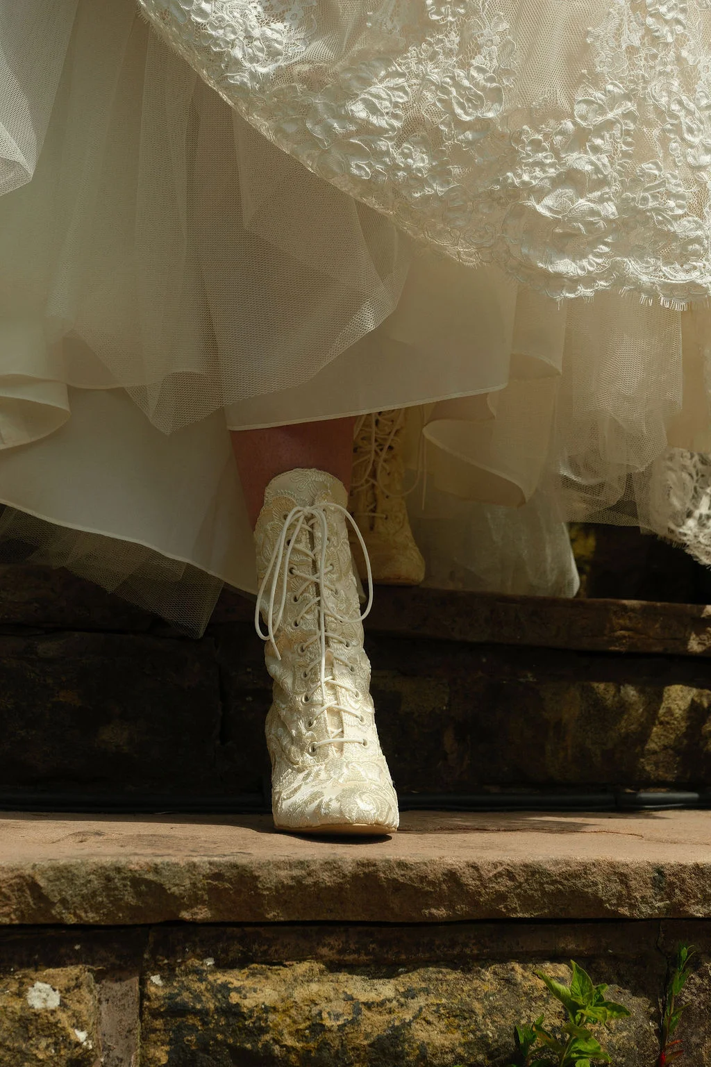 A detailed close-up shot of a bride's ornate lace-up ankle boots peeking out beneath a layered tulle and lace wedding dress as she steps down stone steps.