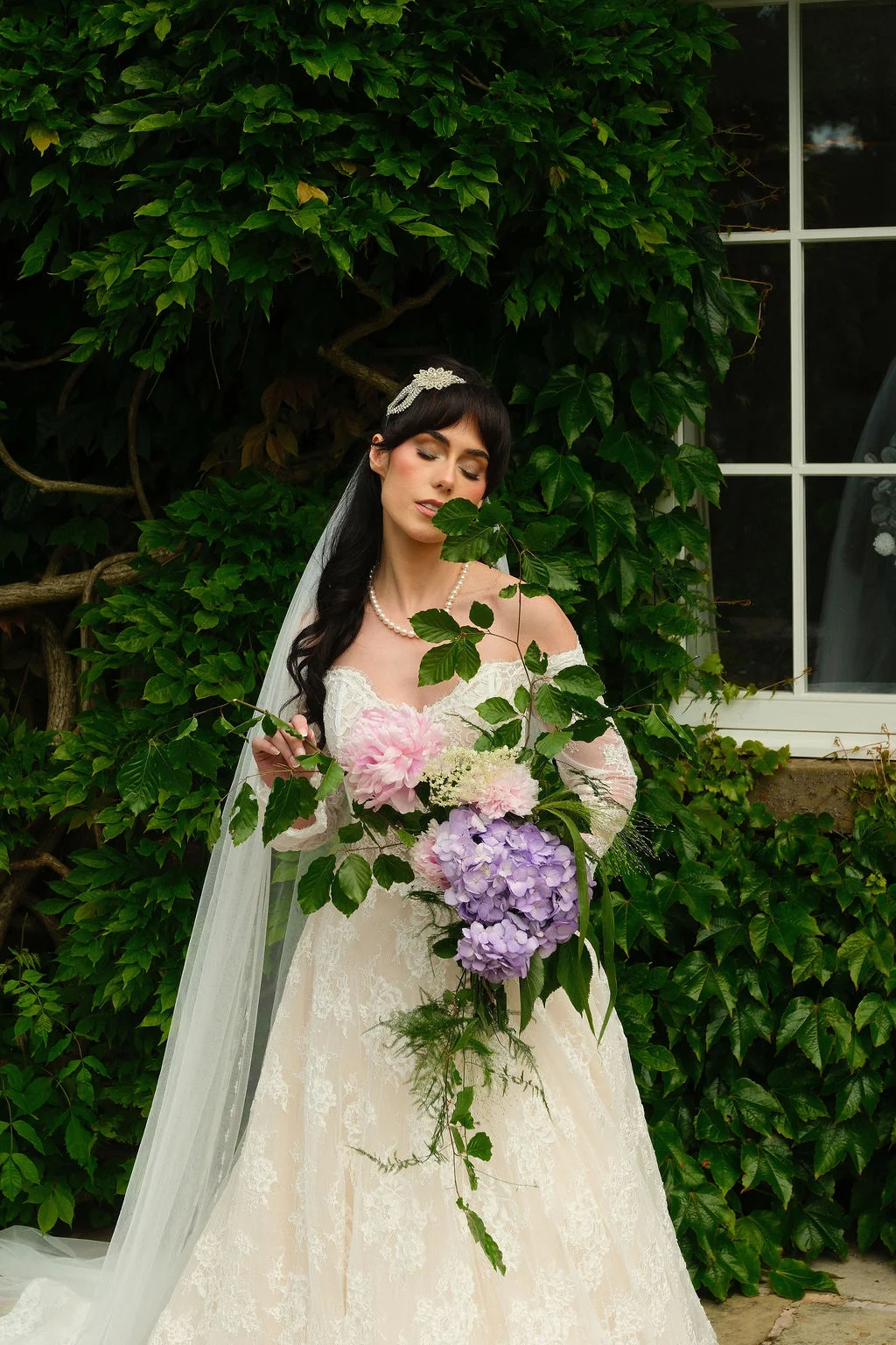 A stunning bride with dark hair and a pearl necklace poses outdoors against an ivy-covered wall, holding a cascading bouquet of pink peonies and purple hydrangeas.